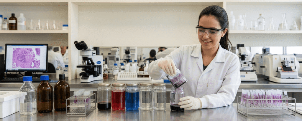 Histotecnologista no laboratório mergulhando suporte de lâminas microscópicas em frasco de coloração.